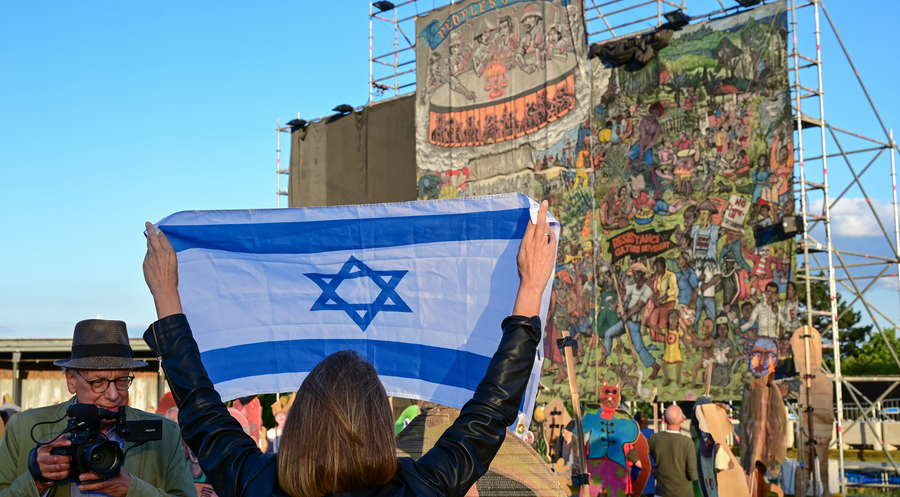 Eine Frau mit israelischer Flagge steht vor dem teilweise verhülltem Großgemälde mit den umstrittenen Figuren des Kollektivs Taring Padi. Foto: Picture Alliance/Uwe Zucchi