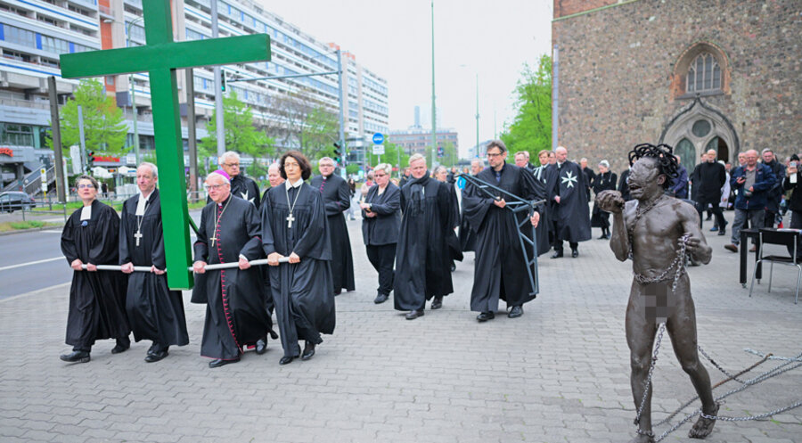 Die Kirchenleiter tragen bei der Karfreitagsprozession ein grünes Kreuz, während ein nackter Künstler in Ketten und Schlamm daneben steht. Foto: picture-alliance/dpa|Sebastian Gollnow